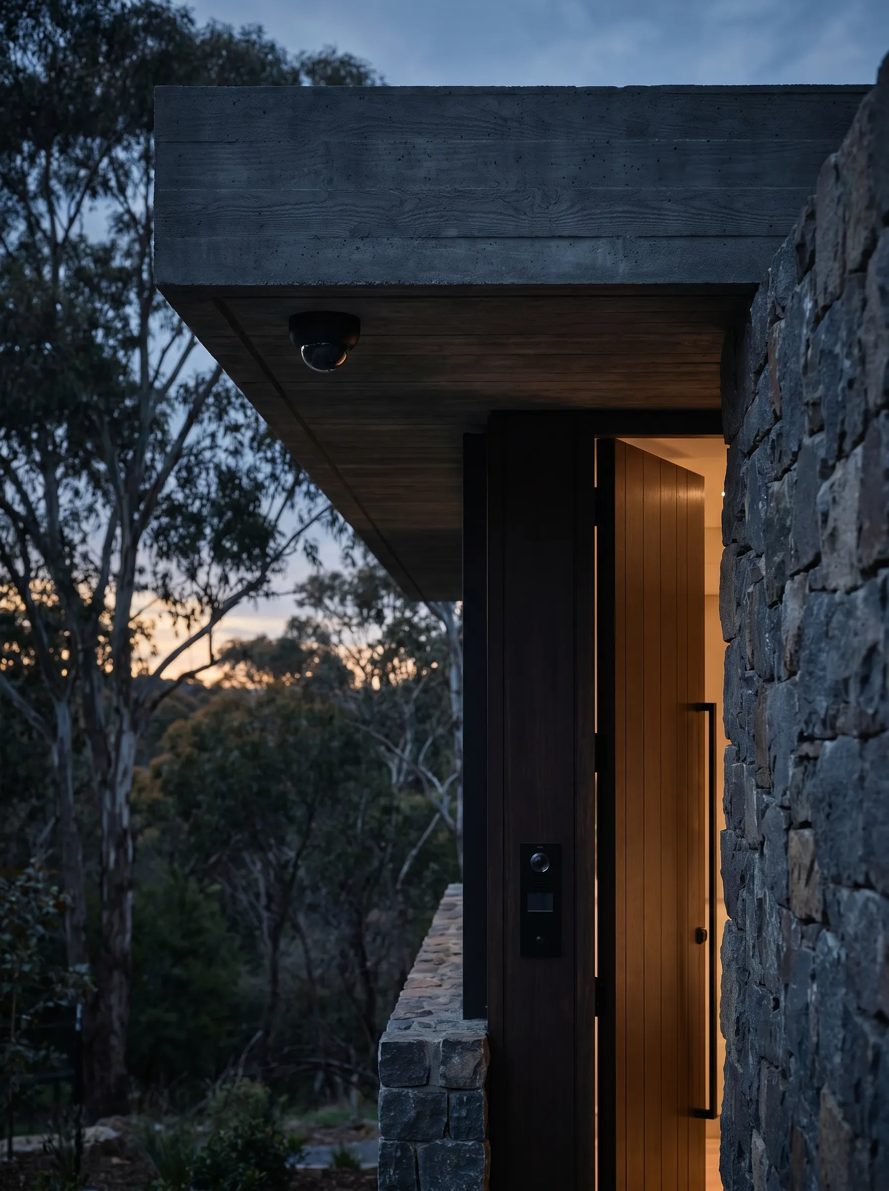 A premium Canberra bushland home entrance at dusk — discreet dome camera on the concrete soffit and an intercom keypad beside the open timber door, gum trees beyond